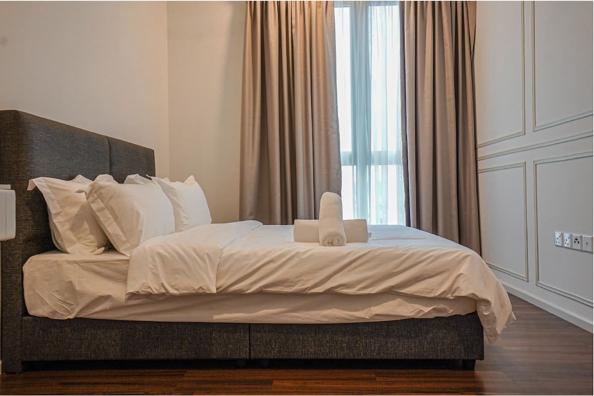 A neatly made bed with white linens and a rolled towel, a dark headboard, and a window with sheer curtains.