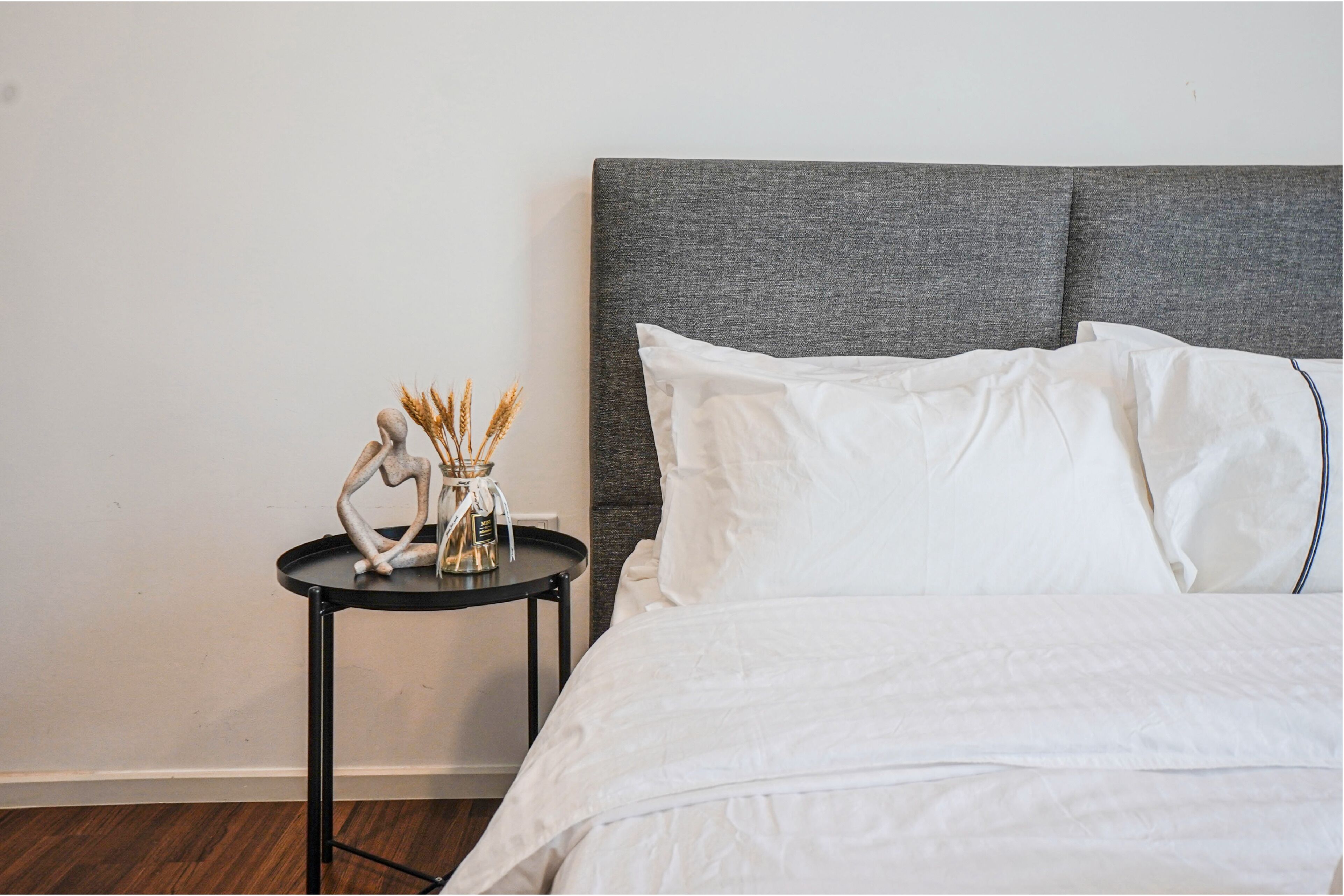 Un lit bien fait, avec du linge de lit blanc, une tête de lit grise et une petite table sur laquelle se trouvent un vase et quelques objets décoratifs.