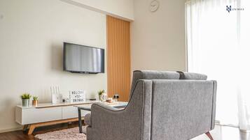 A modern living room with a grey sofa, a flat-screen TV mounted on the wall, a wooden slat partition, and a small table with decorative items.