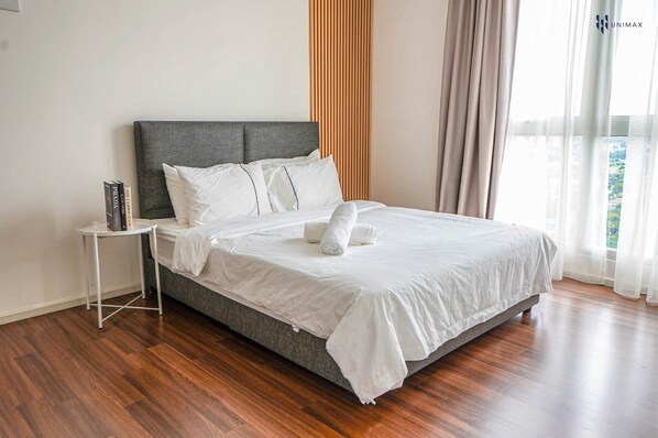 A neatly made bed with white linens and pillows, a small white side table with a book, and a view of greenery through sheer curtains.