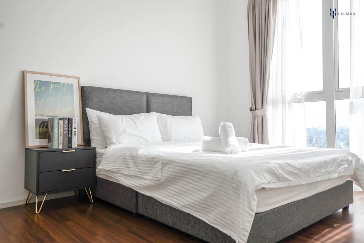 A modern bedroom with a grey headboard, white bedding, a bedside table with books, and a large window with curtains.