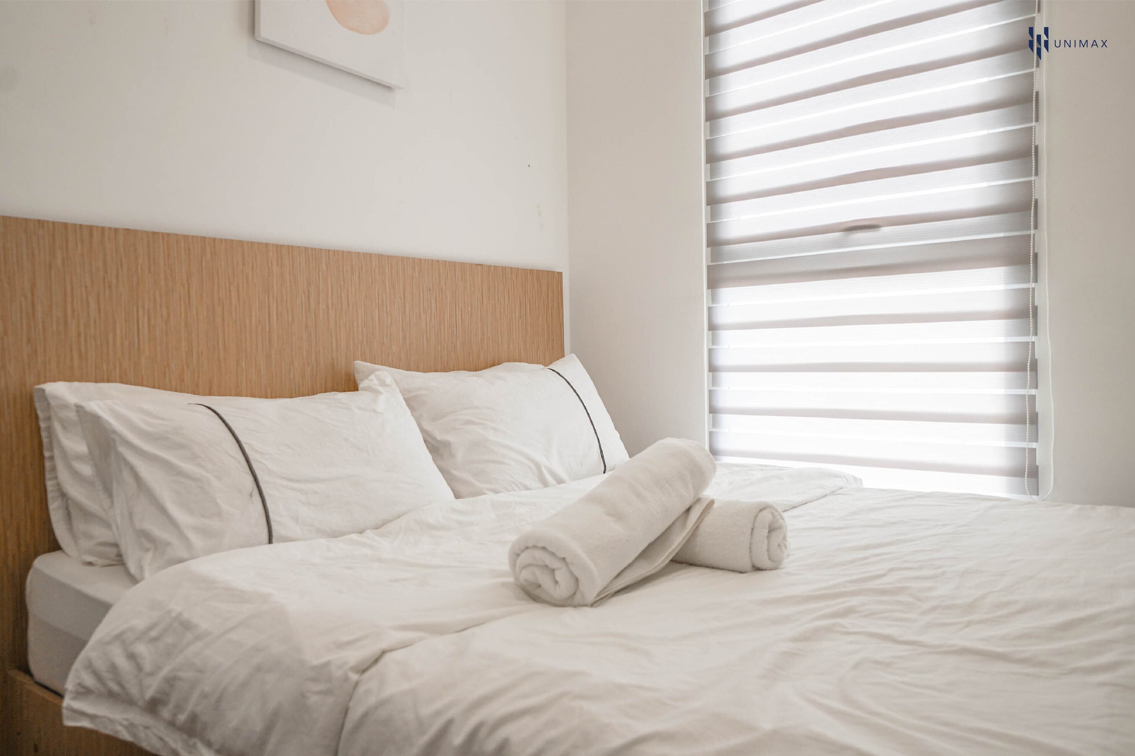A neatly made bed with white linens and a rolled towel, a wooden headboard, and a window with blinds.