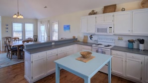 Een ruime keuken met witte kastjes, een centraal eiland en een eethoek met houten stoelen.