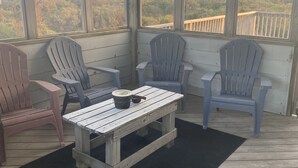 Een overdekt buitengebied met houten tafels en Adirondack-stoelen, met uitzicht op een bebost landschap.
