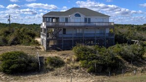 Een twee verdiepingen tellend huis aan het strand met een balkon, omgeven door begroeiing en een heldere lucht.