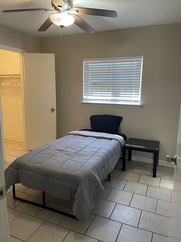 A bedroom with a bed, a nightstand, a window with blinds, a ceiling fan, and a door.