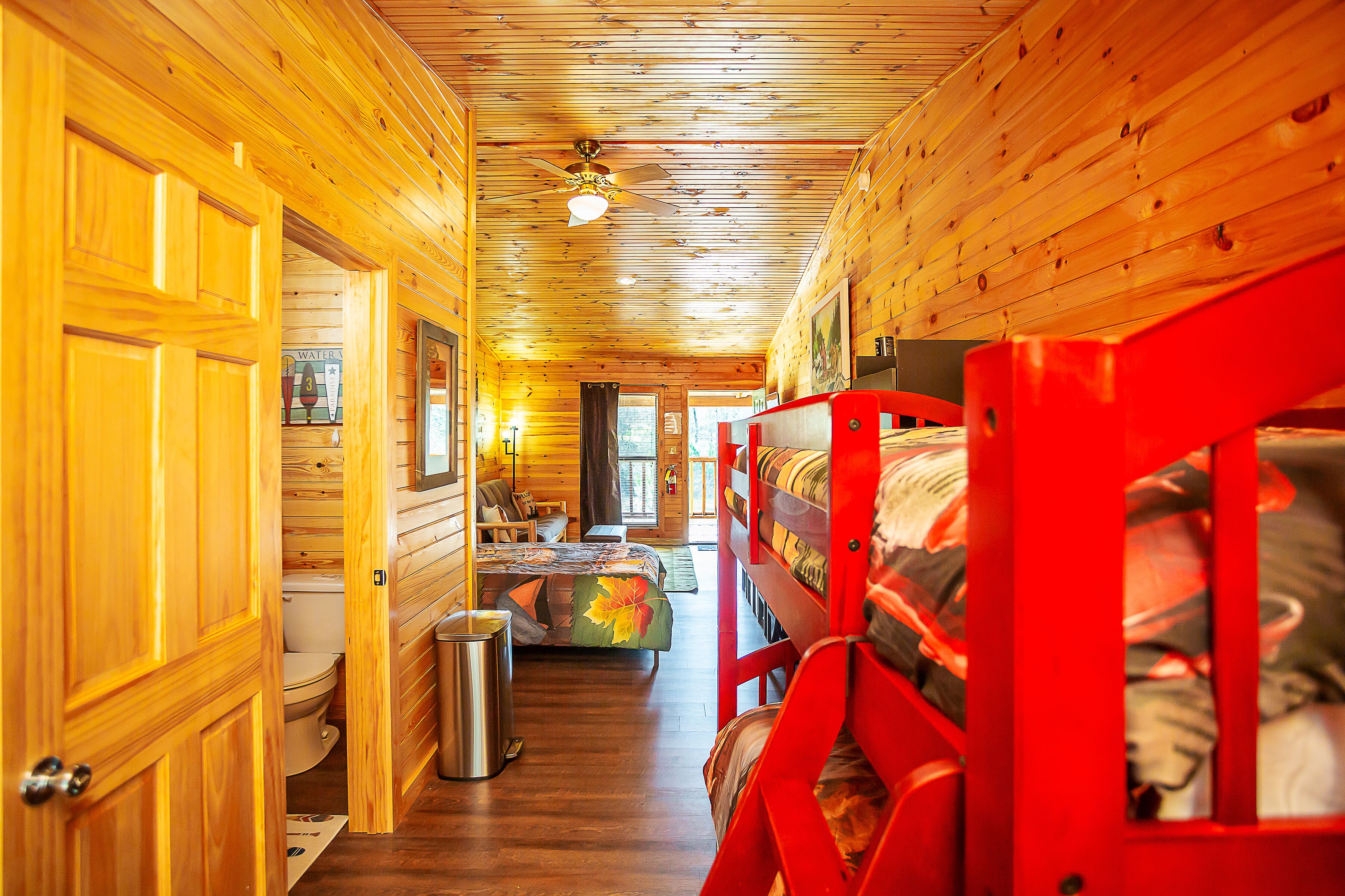 A cabin interior with wooden walls, a ceiling fan, a bunk bed, a bathroom with a toilet, and a trash can.