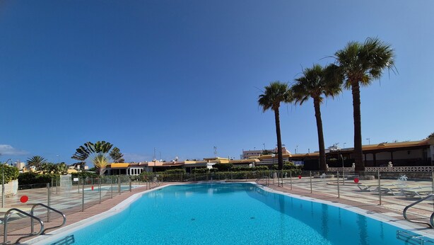 A clear blue swimming pool with a tiled deck, surrounded by palm trees and a building with a yellow roof in the background. - Casa Dailos - 2 bedrooms with washing machine and wifi (Playa Del Ingles)