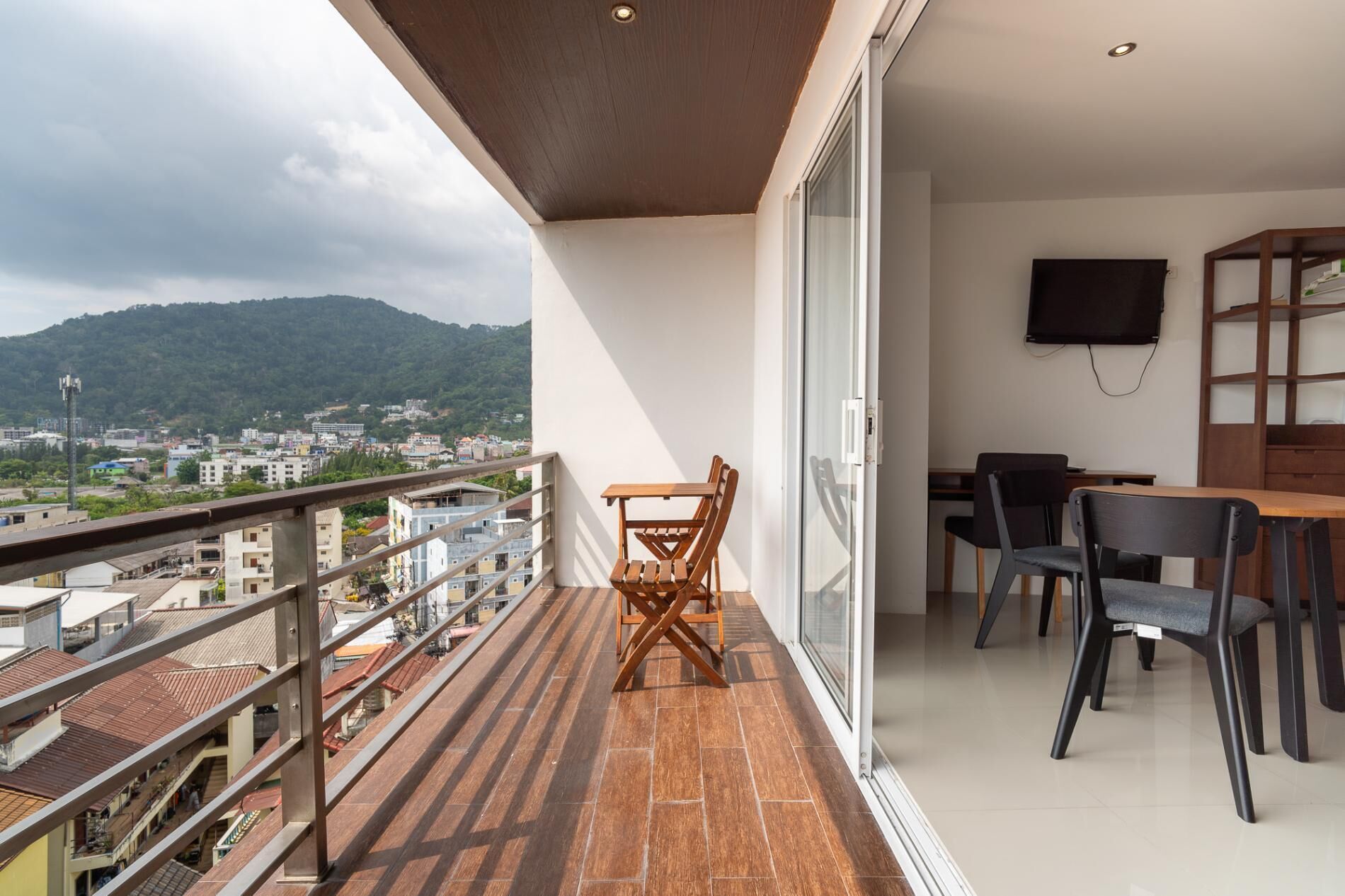 Een balkon met een houten tafel en stoelen, een aan de muur bevestigde televisie en uitzicht over de stad.