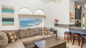 A living room with a large window overlooking a beach, a sofa, a coffee table, and a kitchen area with bar stools. - Mykonos Bay Resort at Zippers Surf Break (San José del Cabo)