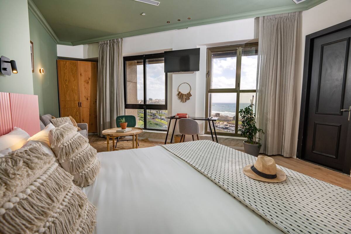 A bedroom with a large bed, a desk, a chair, a TV, and a view of the sea.