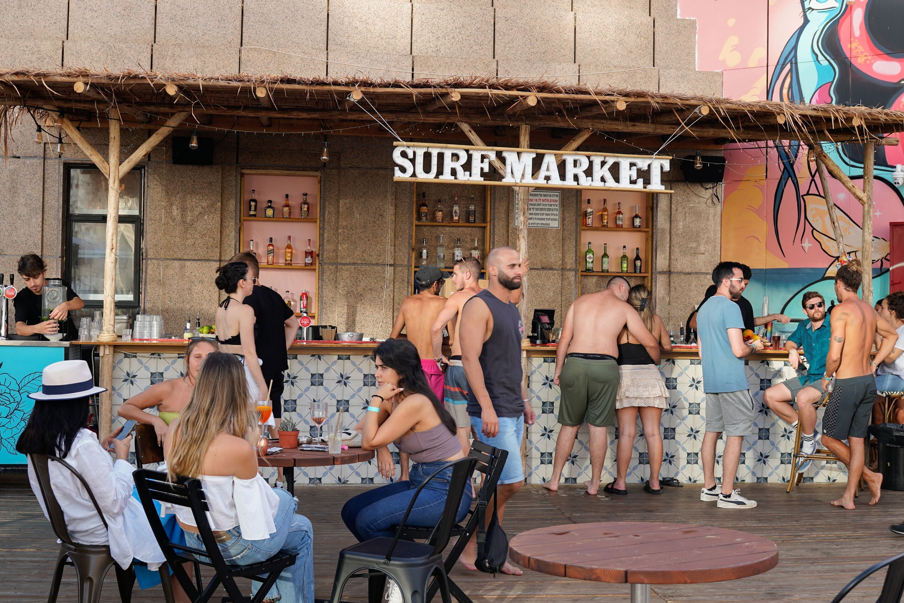 Ett utomhusområde med bar, ett skylt med texten ”Surf Market”, träbord och stolar.