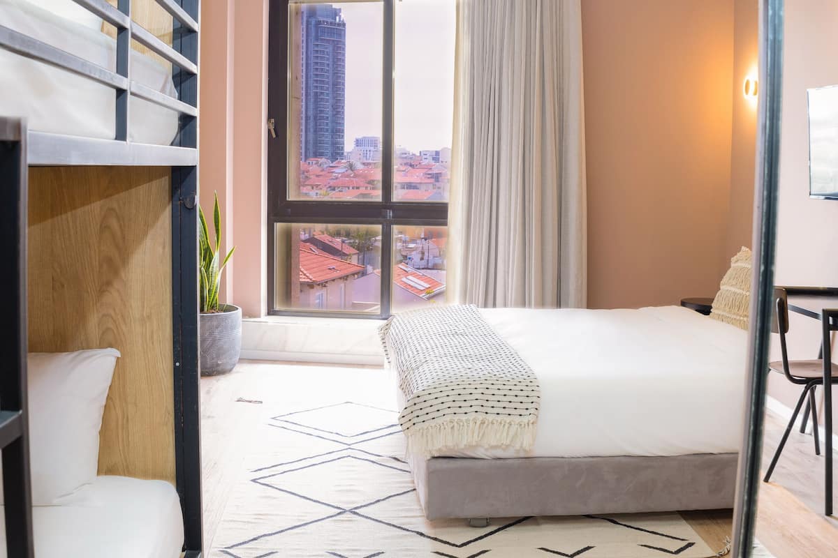 A hotel room with a bed, a chair, a potted plant, and a window offering a city view.