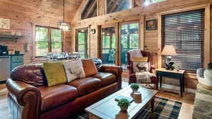 A wooden cabin interior with a leather couch, a dining area, and a window with blinds. - Hot Tub/Close2Downtown/Nearby Fishing Pond (Sevierville)