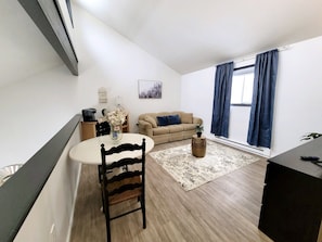 A living room with a sofa, a dining table with a chair, a small rug, and a window with curtains.