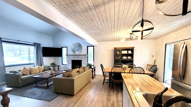 A modern living room with a fireplace, a dining area with a wooden table and chairs, and a kitchen with a stainless steel refrigerator. - ZenNature with hottub & pet friendly (Tobyhanna)