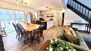 A modern kitchen with a central island, a dining area with a wooden table and chairs, and a living area with a sofa and a staircase. - ZenNature with hottub & pet friendly (Tobyhanna)