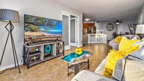 A living room with a TV, a coffee table, and a sofa. There is a kitchen in the background. - Kauhale Makai - Penthouse - Central AC -Oceanfront Unit - Ocean View! (Kihei)