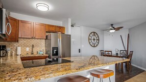 A modern kitchen with granite countertops, wooden cabinets, a built-in refrigerator, and a dining area with a table and chairs. - Kauhale Makai - Penthouse - Central AC -Oceanfront Unit - Ocean View! (Kihei)