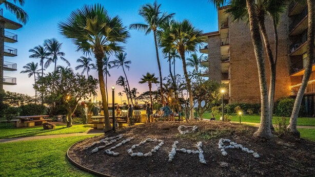 A landscaped area with palm trees and a circular garden bed with the word "LOHA" spelled out. - Kauhale Makai - Penthouse - Central AC -Oceanfront Unit - Ocean View! (Kihei)
