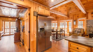 Interior de una cabaña de madera con un refrigerador de acero inoxidable, armarios de madera, una zona de comedor con mesa y sillas, y una chimenea acogedora.