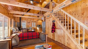 Interior de una cabaña de madera con sofá, una mesa de comedor, una escalera y un letrero que indica un sendero para caminatas.