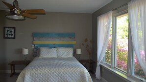 Un dormitorio con cama, dos mesitas de noche, ventilador de techo, ventana con cortinas y vistas a flores fuera.
