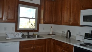 Una cocina con armarios de madera, encimera blanca, pileta, microondas, lavavajillas y ventana con vista a follaje.
