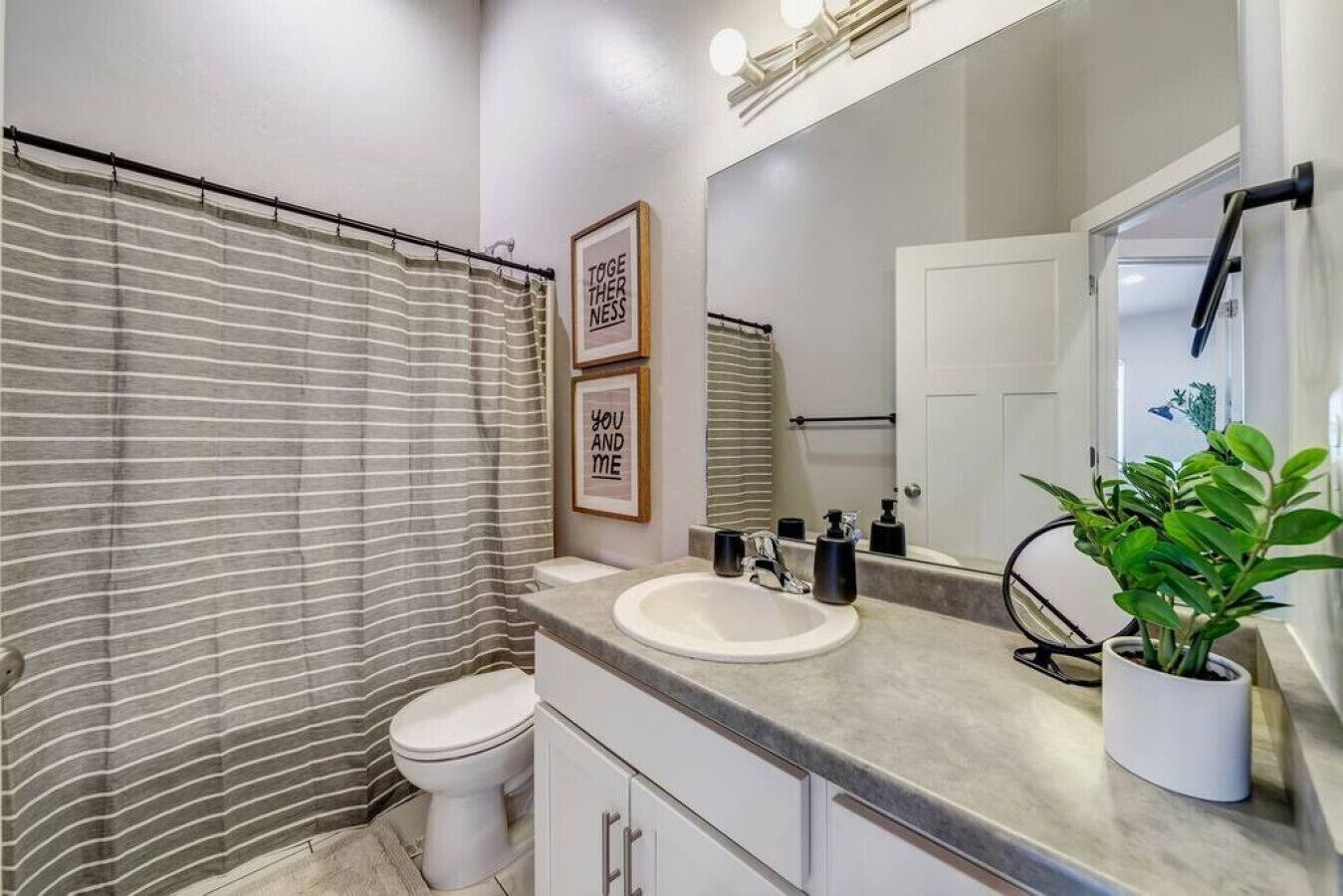 A modern bathroom with a striped shower curtain, a toilet, a sink with a mirror above it, and two framed motivational quotes on the wall.
