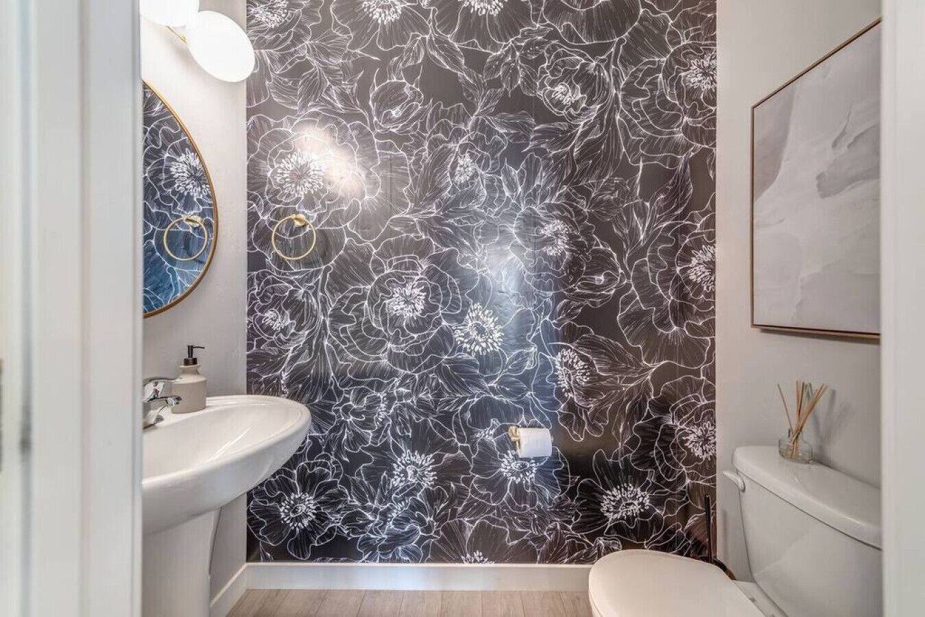 Une salle de bain avec un papier peint à motifs floraux, un lavabo, des toilettes et une œuvre d’art encadrée accrochée au mur.