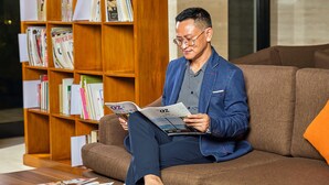 A person sitting on a brown sofa reading a magazine in a room with a wooden bookshelf filled with books and magazines.