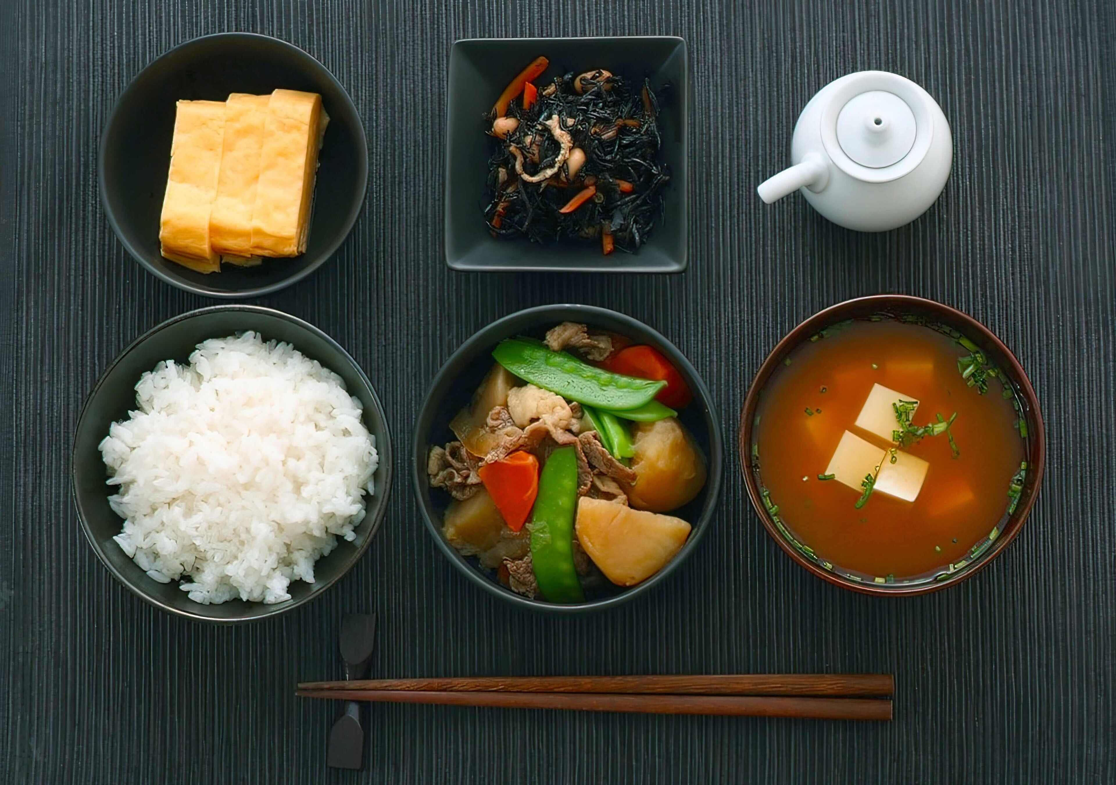 A meal setup with tofu, seaweed, rice, a vegetable stir-fry, and a soup with tofu, arranged on a dark surface.