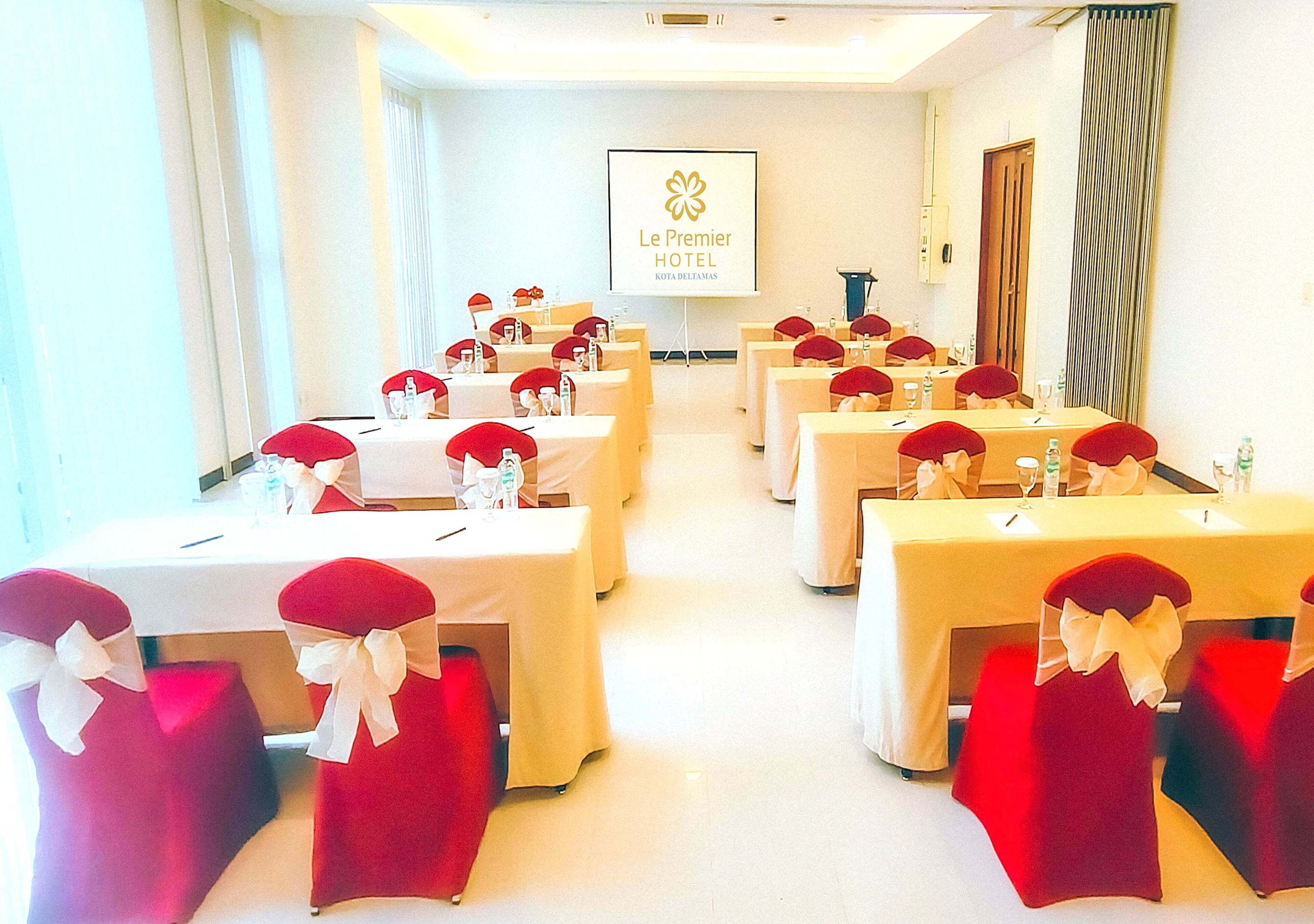 A conference room with tables set for a meeting, each with a red chair and white tablecloth. A projector screen displays the hotel name.