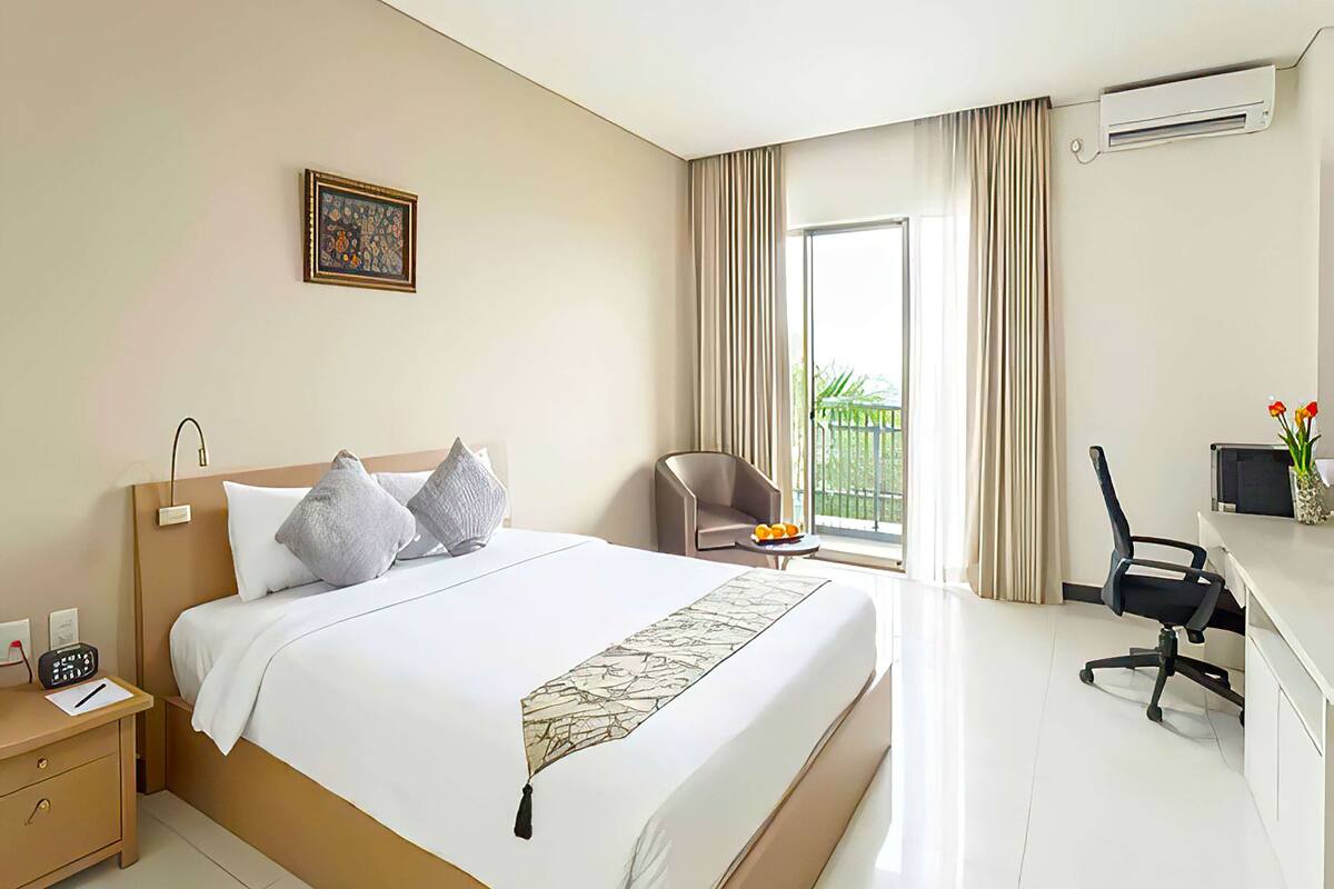 Una habitación de hotel con una cama grande, un escritorio con una silla, una mesita con fruta y una ventana con cortinas.