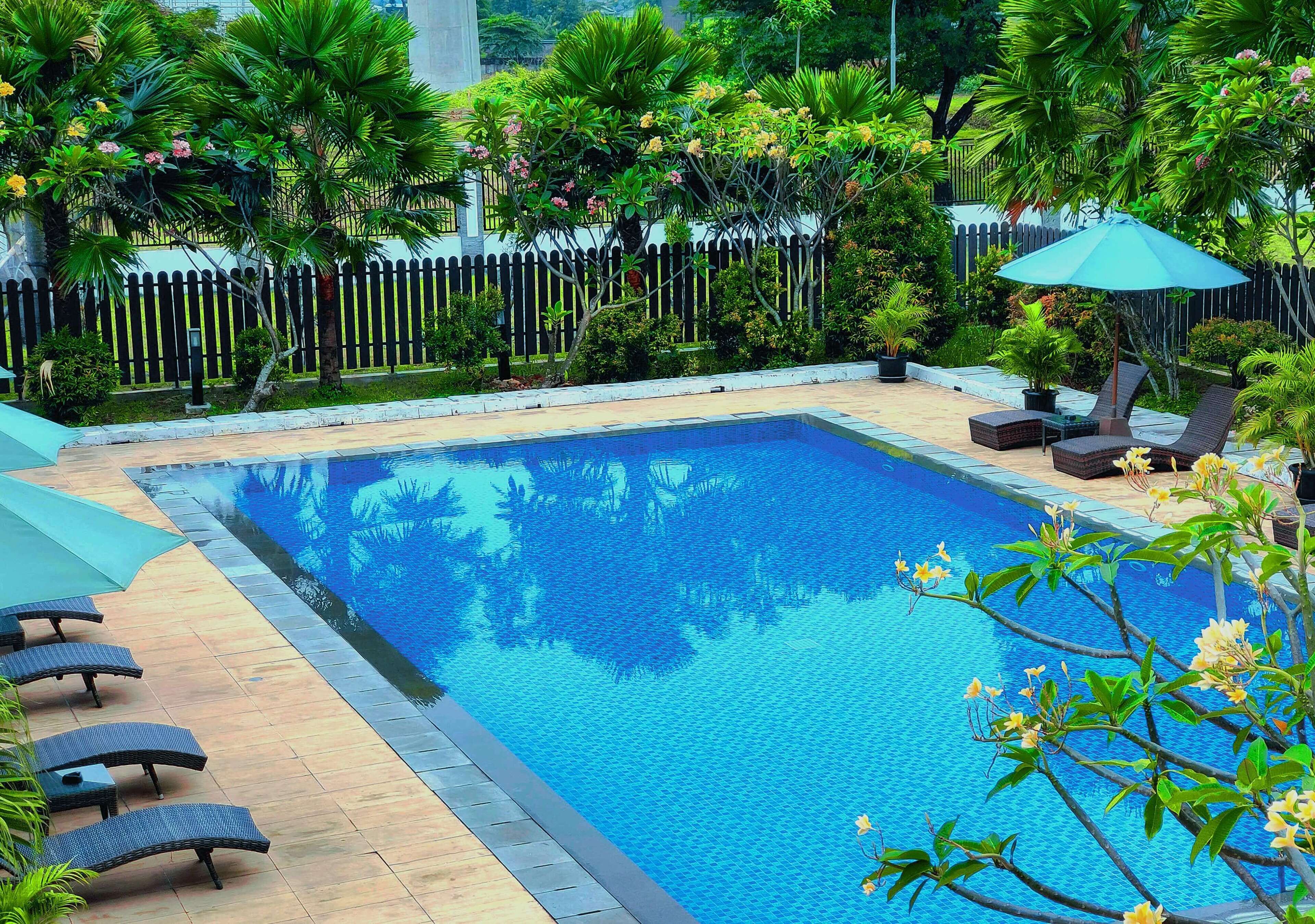 A rectangular swimming pool with a wooden deck, surrounded by lush greenery and a wooden fence.