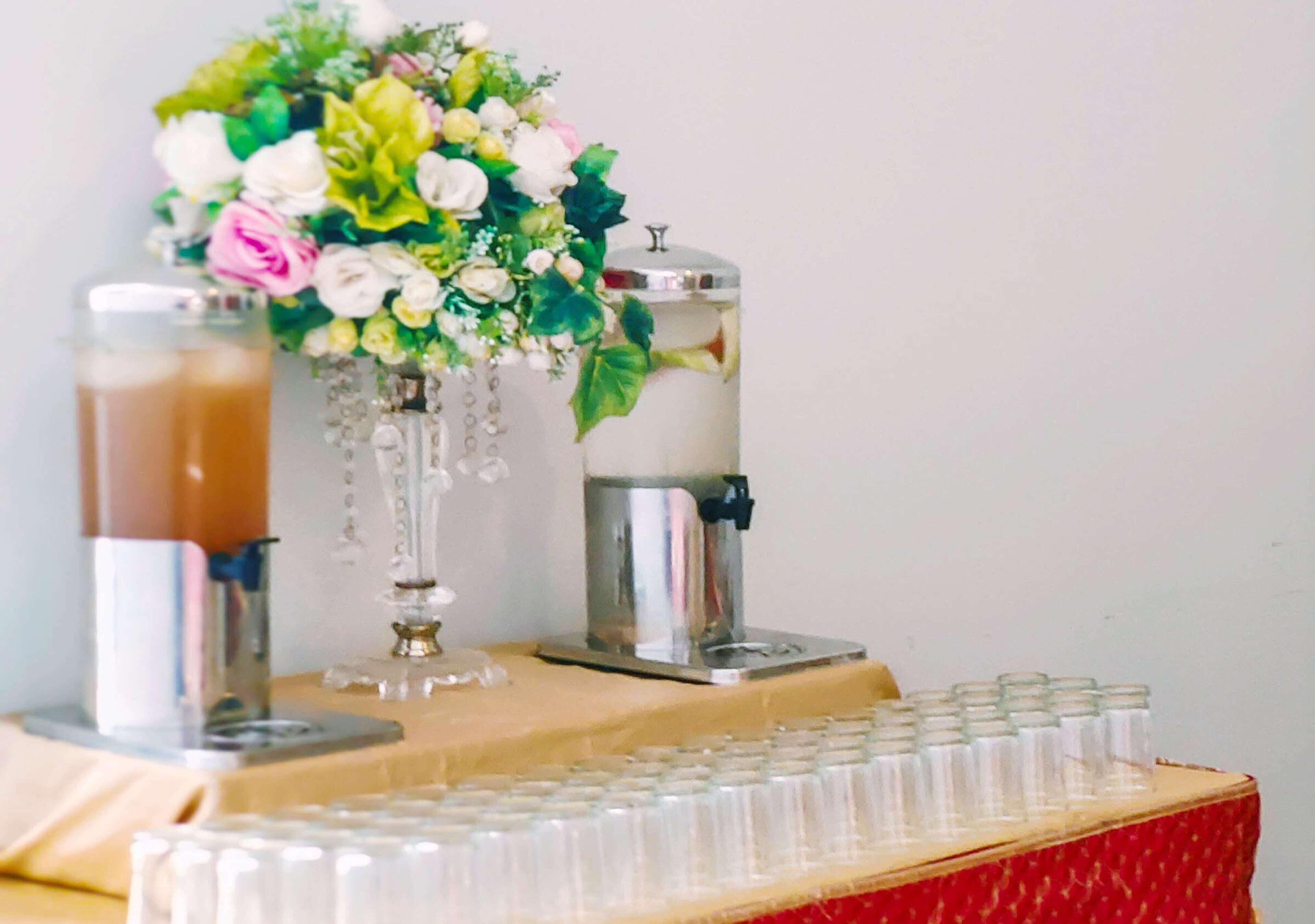 A table with a floral arrangement, two cylindrical containers, and a row of clear plastic cups.