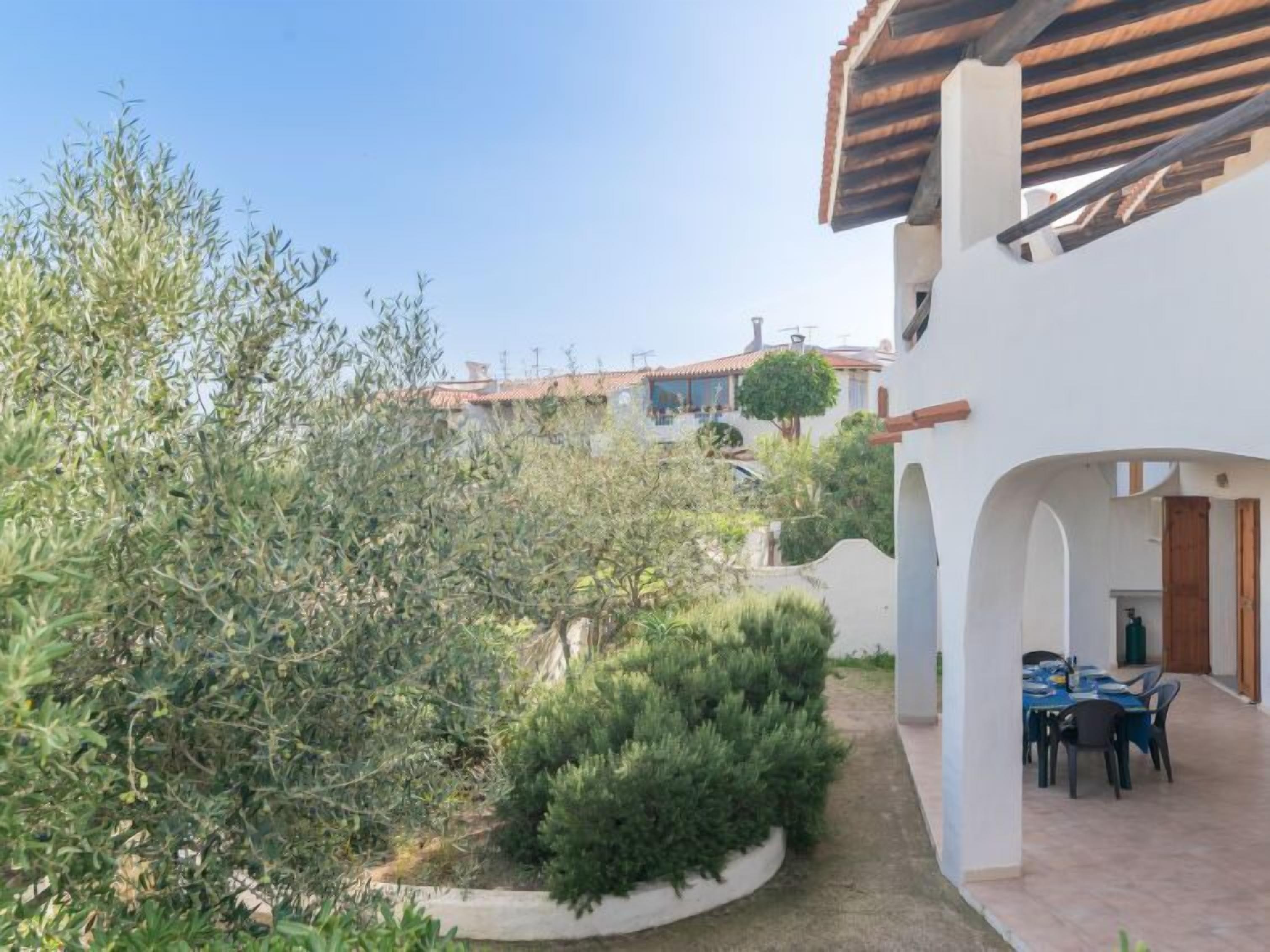 A courtyard with a dining area, surrounded by olive trees and a white building with a tiled roof.