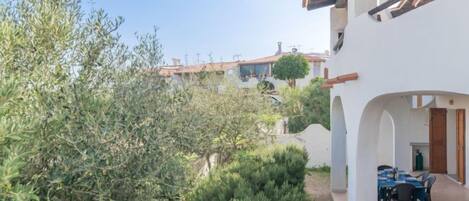 A courtyard with a dining area, surrounded by olive trees and a white building with a tiled roof.