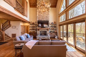 A spacious living room with a stone fireplace, a large television, and a cozy seating area. - Antler Ridge - Eagles Nest home with big views, 3 suites, pool table, hot tub, EV charger (Elk Park)
