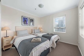A bedroom with two beds, a nightstand with a lamp, a window with blinds, and framed pictures on the wall.