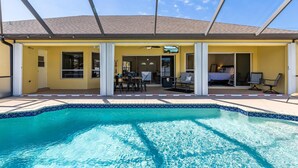 A pool area with a clear blue pool, a covered patio with chairs, and a building with a yellow exterior. - Gorgeous heated pool home - Villa Palm Tree - Roelens Vacations (Cape Coral)