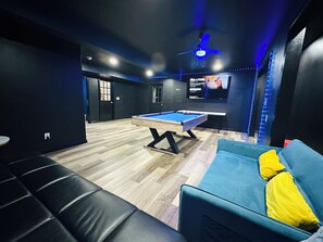 Ein moderner Raum mit Pooltisch, einem blauen Sofa mit gelben Kissen und einem Deckenventilator.