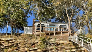 Una cabaña rústica con terraza de madera y pared de piedra, rodeada de árboles y terreno rocoso.