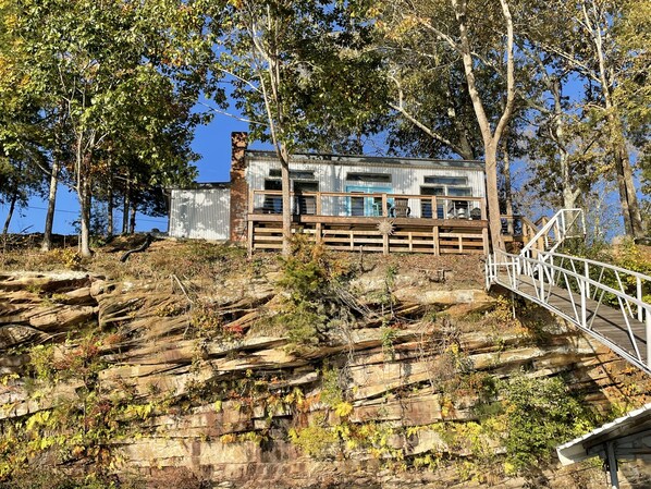A rustic cabin with a wooden deck and a stone wall, surrounded by trees and rocky terrain.