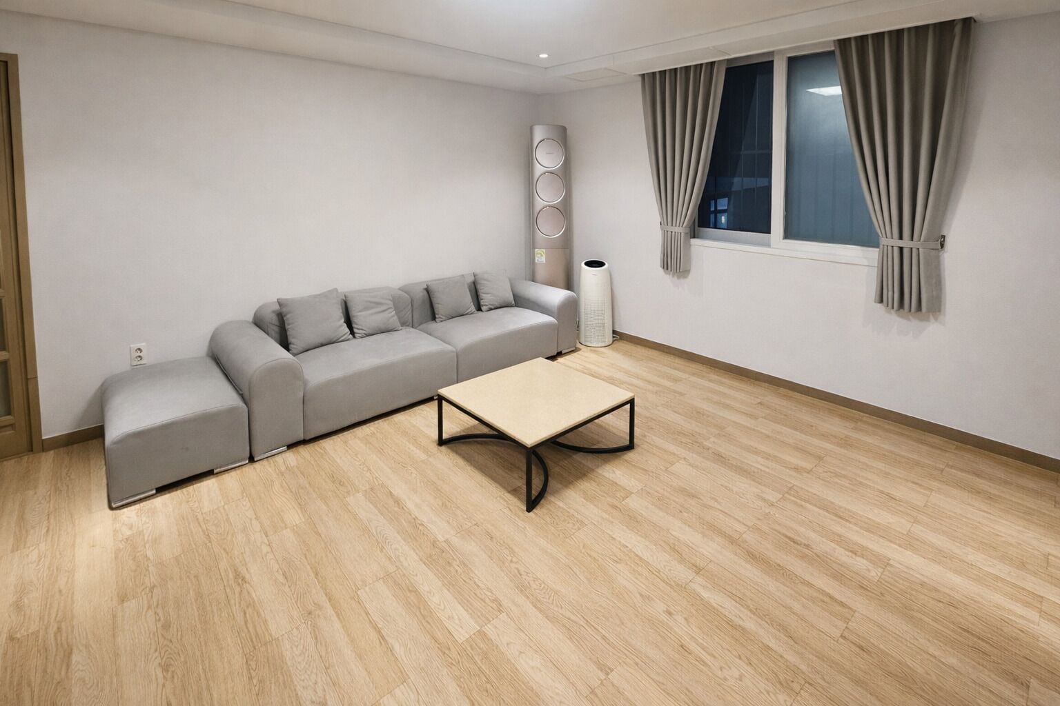 A modern living room with a grey sofa, a small ottoman, a coffee table, a white radiator, and a window with curtains.