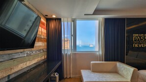 Habitación de hotel con un televisor grande, un sofá, un ventanal con vistas a un puente y una frase inspiradora en la pared.