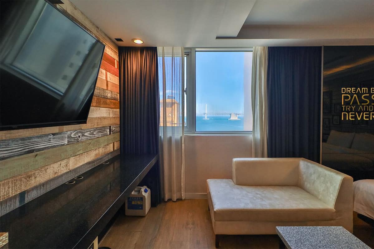 A hotel room with a large TV, a sofa, a window with a view of a bridge, and a motivational quote on the wall.