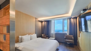 Habitación de hotel con una cama grande, una mesita, dos sillas y vista al mar a través de amplios ventanales.