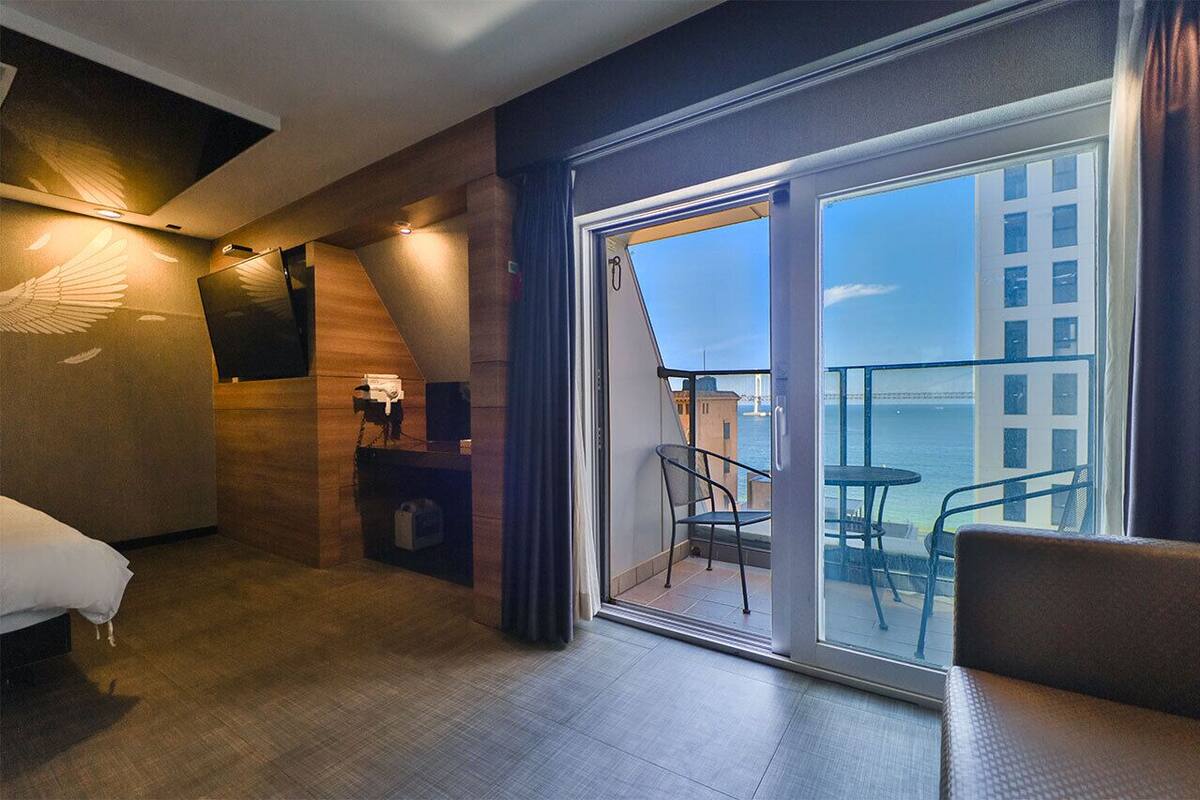 A hotel room with a bed, a desk with a lamp, a chair, and a balcony with a view of the city.