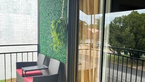 Un balcón con un sofá de mimbre y una mesa, un muro verde con follaje y vistas a una carretera y árboles.